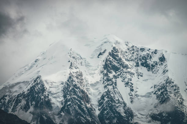 istock Atmospheric mountains landscape with big snowy mountain top with snow cornices in low clouds. Awesome minimal scenery with snow-white high pinnacle in overcast weather. Giant mountain wall in clouds. 1313106970