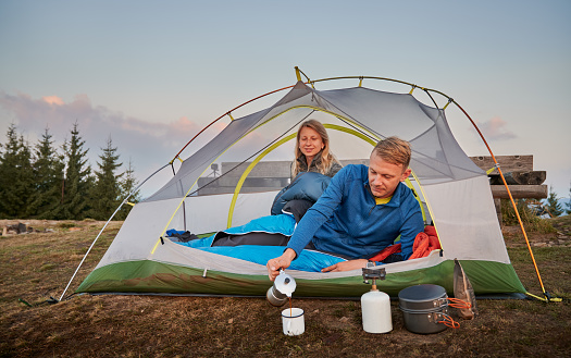 Husband, after waking up in the morning, lying in white tent, pouring freshly brewed hot coffee into metal cup for his wife. Active rest in camp in the mountains. Concept of hiking and camping.