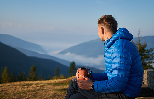 Hiking in the mountains. Guy sitting outdoors on mountain meadow at sunny morning, looking at very beautiful misty mountain hills. Active trekking recreation.