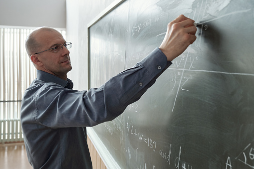 Mature professor of university in smart casualwear standing in front of blackboard at lesson in auditorium and writing down formula