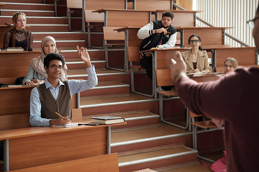 Young successful student in eyeglasses and casualwear raising hand while sitting by desk on the first row and looking at professor at lesson