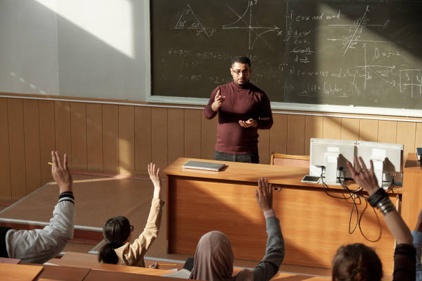 Middle aged teacher in casualwear standing by blackboard in lecture hall and looking at students Middle aged confident teacher in casualwear standing by blackboard in lecture hall and looking at group of students ready to answer lecture hall stock pictures, royalty-free photos & images