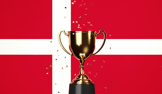 Star shaped gold confetti falling onto a gold cup sitting over Danish flag background. Horizontal composition with copy space. Front view. Championship concept.