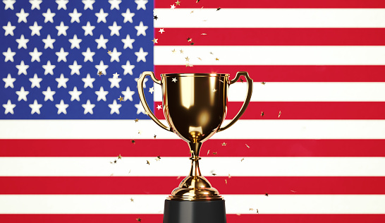 Star shaped gold confetti falling onto a gold cup sitting over American flag background. Horizontal composition with copy space. Front view. Championship concept.