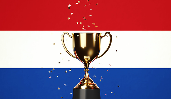 Star shaped gold confetti falling onto a gold cup sitting over Dutch flag background. Horizontal composition with copy space. Front view. Championship concept.