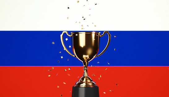 Star shaped gold confetti falling onto a gold cup sitting over Russian flag background. Horizontal composition with copy space. Front view. Championship concept.