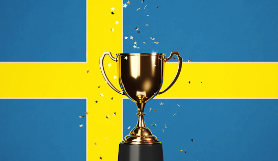 Star shaped gold confetti falling onto a gold cup sitting over Swedish flag background. Horizontal composition with copy space. Front view. Championship concept.