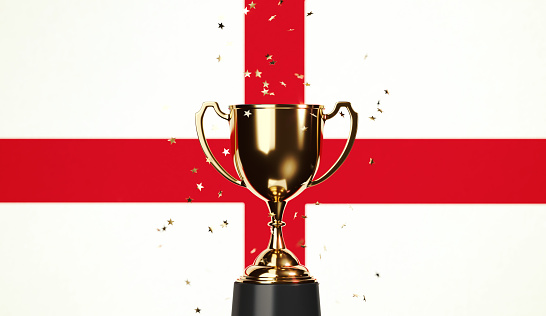 Star shaped gold confetti falling onto a gold cup sitting over English flag background. Horizontal composition with copy space. Front view. Championship concept.
