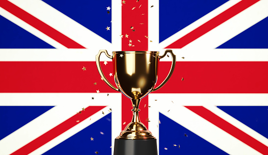 Star shaped gold confetti falling onto a gold cup sitting over British flag background. Horizontal composition with copy space. Front view. Championship concept.