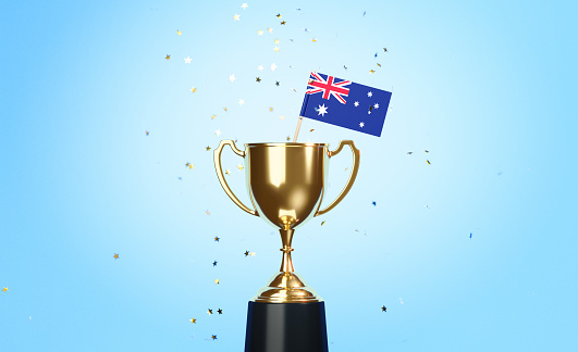 Star shaped gold confetti falling onto a gold cup in which a tiny Australian flag sitting before blue background. Horizontal composition with copy space. Front view. Championship concept.