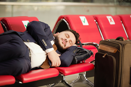 Tired middle aged businessman in formalwear lying on row of red leather seats and napping in lounge of modern airport while waiting for flight