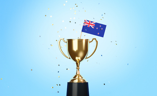 Star shaped gold confetti falling onto a gold cup in which a tiny New Zealand flag sitting before blue background. Horizontal composition with copy space. Front view. Championship concept.