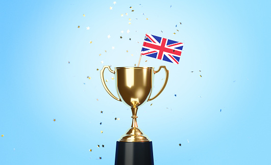 Star shaped gold confetti falling onto a gold cup in which a tiny British flag sitting before blue background. Horizontal composition with copy space. Front view. Championship concept.