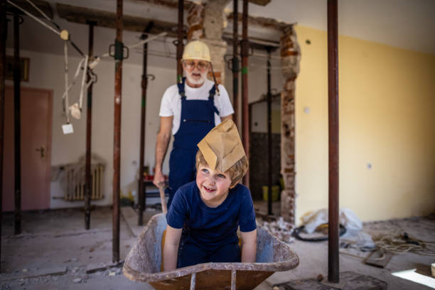 Photo of a grandfather and his grandson working together on a home reconstruction area.Grandfather pushed his grandson in a wheelbarrow on a construction cite.