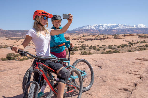 Female mountain bikers follow Slickrock trail in the morning They take photos of distant scene on cell phone, snowy mountain ranges in the distance slickrock trail stock pictures, royalty-free photos & images