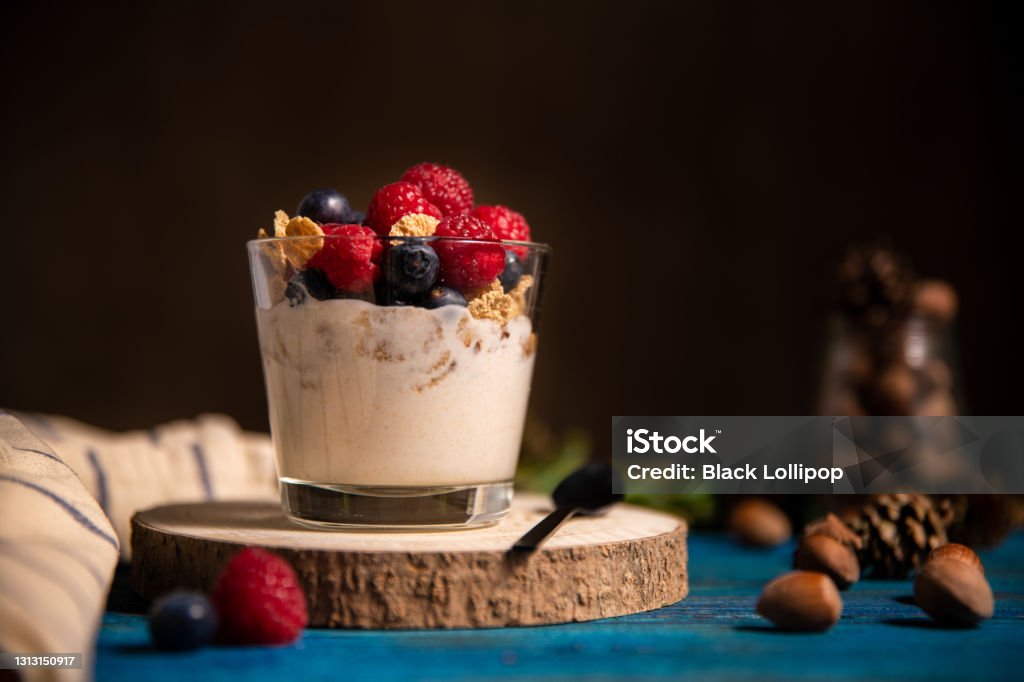 Granola with yogurt and berries for healthy breakfast on a wooden table. Dark style photography. Glass of homemade granola with yogurt and fresh raspberries, hazelnut and blueberries on blue wooden background. Fitness breakfast. Space for text on dark background. Yogurt Stock Photo