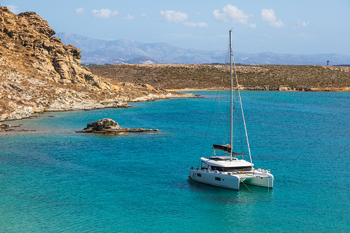 Large white sailboat anchored in a small bay with blue turquoise water, retraced sail, aerial view, village at the waterfront, mountain range and clear sky in background