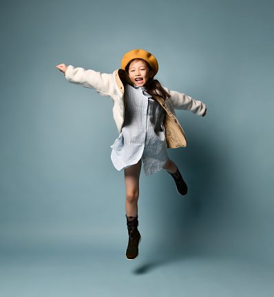 Little asian girl in double sided jacket, shirt dress, brown beret, boots. She laughing out loud, jumping up against blue studio background. Childhood, fashion, hipster style. Full length, copy space