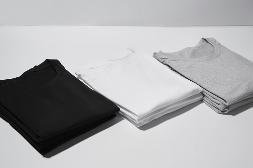 Three folded monochrome t shirts gray, black and white isolated over gray background. Clothes, fashion, retail concept. Horizontal shot