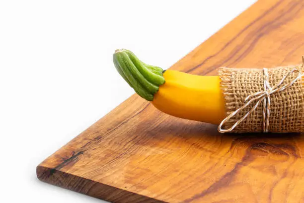 Photo of Single Summer Squash Zucchini, Yellow Colored Gourd Also Known As Courgette, Baby Marrow, Indian Jugni Sabzi Or Jugnu Petha Sabji Tied In Burlap Cloth On Cutting Board. White Background And Copy Space