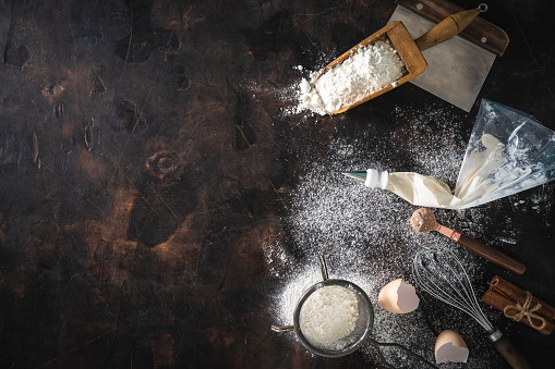Bakery background with flour, eggs, sieve, whisk, serving scoop, cinnamon, pastry bag  and Bench Scraper on dark wood background
