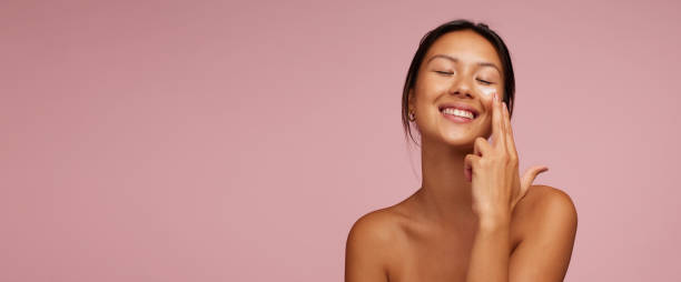 Woman applying skincare product on her face Woman applying skincare product on her face. Asian woman putting cosmetic cream in her face and smiling against pink background. face cream stock pictures, royalty-free photos & images
