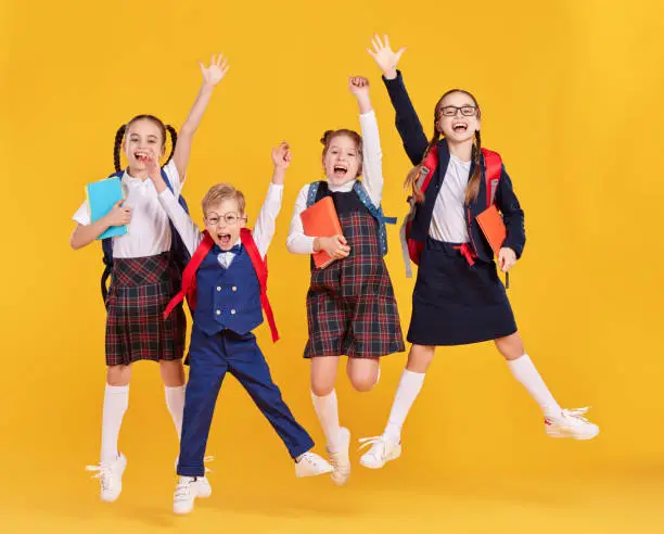 Group of funny excited classmates girls and boy in school outfits with backpacks and copybooks having fun and jumping on yellow background