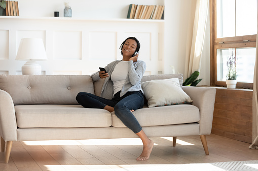 Full length relaxed young african american biracial woman resting on comfortable couch, dreaming listening favorite music in wireless headphones, choosing audio tracks in mobile app library at home.