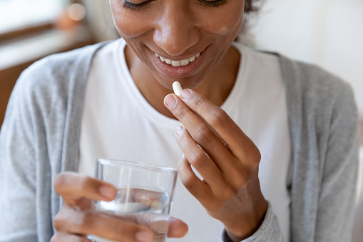 Close up happy young african american young woman taking daily dose of complex healthcare skin, hair and nails omega vitamins drinking glass of fresh pure water, immunity improvement concept.