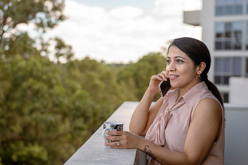 A beautiful woman of Indian descent stands on her apartment patio and talks on the phone while enjoying a cup of coffee. The relaxed woman is enjoying the outdoors and  is staying connected with family and friends while living in the city. Social distancing, mental health, and life balance concepts.