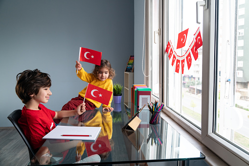 Photo of 8 years old elementary schoolboy and 2,5 years old sister celebrating National Sovereignty And Children's Day at home via video conferencing on digital tablet in new normal. They are sitting on desk next to window. Shot indoor with a full frame mirrorless camera.