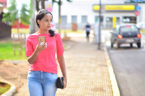 A young Brazilian woman waking on streets of a rural city in the state of Minas Gerais, Brazil, holding a mobile phone and looking for an address on the map. Behind her there is a sidewalk, some shops, trees of a park and some cars and people, all defocussed.
