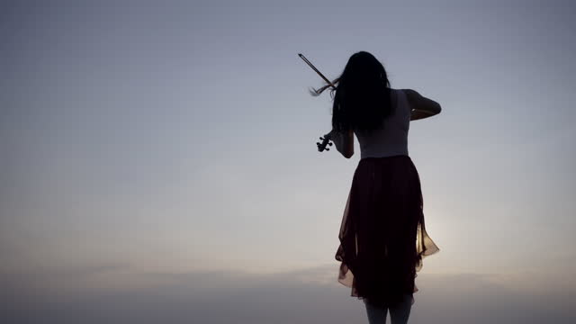 Woman on the mountain playing the violin on sea background at sunset .