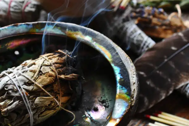 A close up image of a burning white sage smudge stick, healing crystals, and sacred feather.