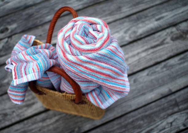 Heading to a day at the beach; beach towels in woven straw tote bag Two colorfully striped beach towels, one rolled, one loose, spilling out of woven straw beach tote with leather handles, on worn, weathered, wooden deck, viewed from above with selective focus on towels straw bag stock pictures, royalty-free photos & images