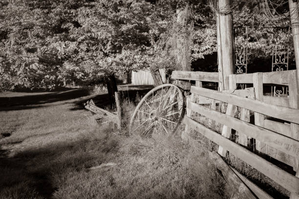 Sepia toned old buildings and wagon wheel among large trees with bright geen and yellow foliage.