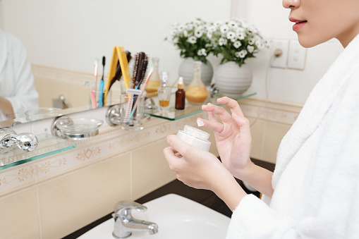 Close-up image of young woman standing at bathroom mirror and applying rich nourishing cream on her face