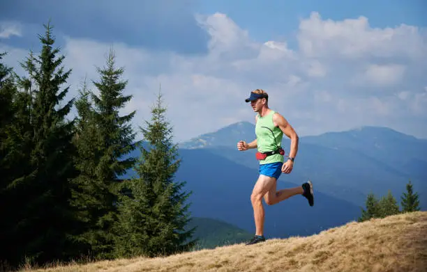 Professional young blond athlete running trail on grassy downhill of mountains in summer sunny day. Beauty mountain landscape on background.