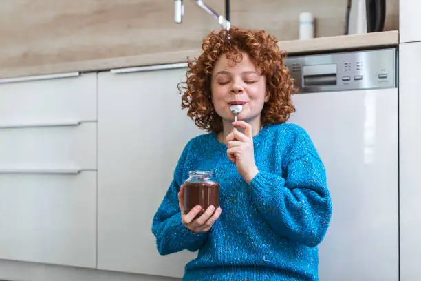 Photo of young woman eating chocolate from a jar while sitting on the wooden kitchen floor. Cute ginger girl indulging cheeky face eating chocolate spread from jar using spoon savoring every mouthful