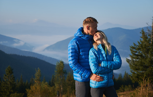 Male traveler hugging smiling woman and kissing her forehead while standing outdoors with blue sky and mountains on background. Happy couple sharing tender kiss while travelling together.