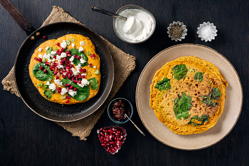 Gluten free recipe for red lentil pancakes; red lentils soaked overnight and blended, chickpea flour, chilli powder, cumin, tumeric, greek yoghurt, spinach leaves, spring onions and fresh coriander.  Served with feta cheese, fresh parsley and pomegranate seeds. Overhead view, horizontal format with some copy space