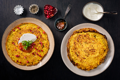 Gluten free recipe for red lentil pancakes; red lentils soaked overnight and blended, chickpea flour, chilli powder, cumin, tumeric, greek yoghurt, spinach leaves, spring onions and fresh coriander.  Served with more yoghurt, fresh parsley and pomegranate seeds and chutney. Overhead view, horizontal format with some copy space