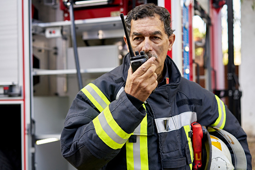 Waist-up view of serious senior man in fire protection suit with helmet under his arm communicating outdoors on portable handheld two-way radio.