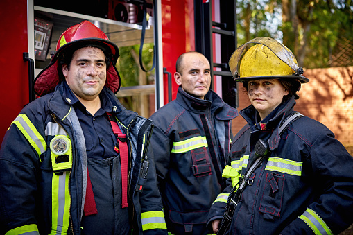 Waist-up view of Caucasian firefighters with sooty faces wearing protection suits and helmets and looking at camera with serious expressions.