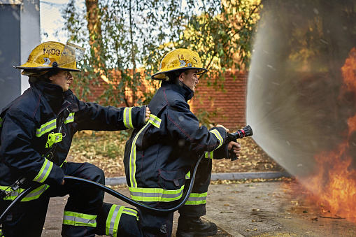 Side view of kneeling female backup firefighter in protection suit and helmet holding the line and steadying shoulder of nozzle man.