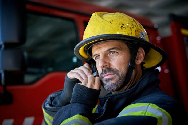 Bearded Firefighter in Mid 40s Putting on Helmet Head and shoulders view of Caucasian man standing in station wearing fire protection suit and looking away from camera while securing chin strap. firefighter photos stock pictures, royalty-free photos & images