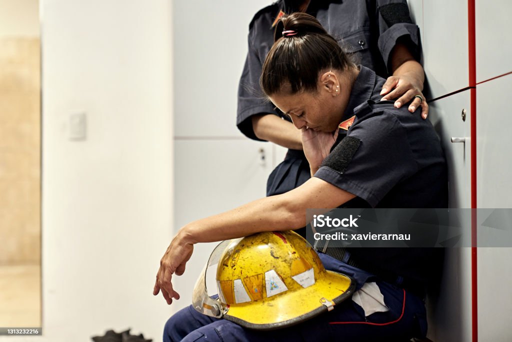 Teammate Consoling Exhausted Female Firefighter Side view of late 30s woman in uniform sitting on bench in fire station locker room with helmet in lap, head down, and eyes closed. Emergency Services Occupation Stock Photo