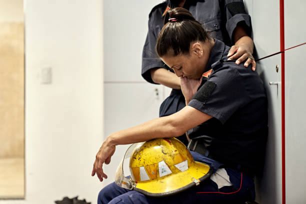 Teammate Consoling Exhausted Female Firefighter Side view of late 30s woman in uniform sitting on bench in fire station locker room with helmet in lap, head down, and eyes closed. hand on shoulder photos stock pictures, royalty-free photos & images