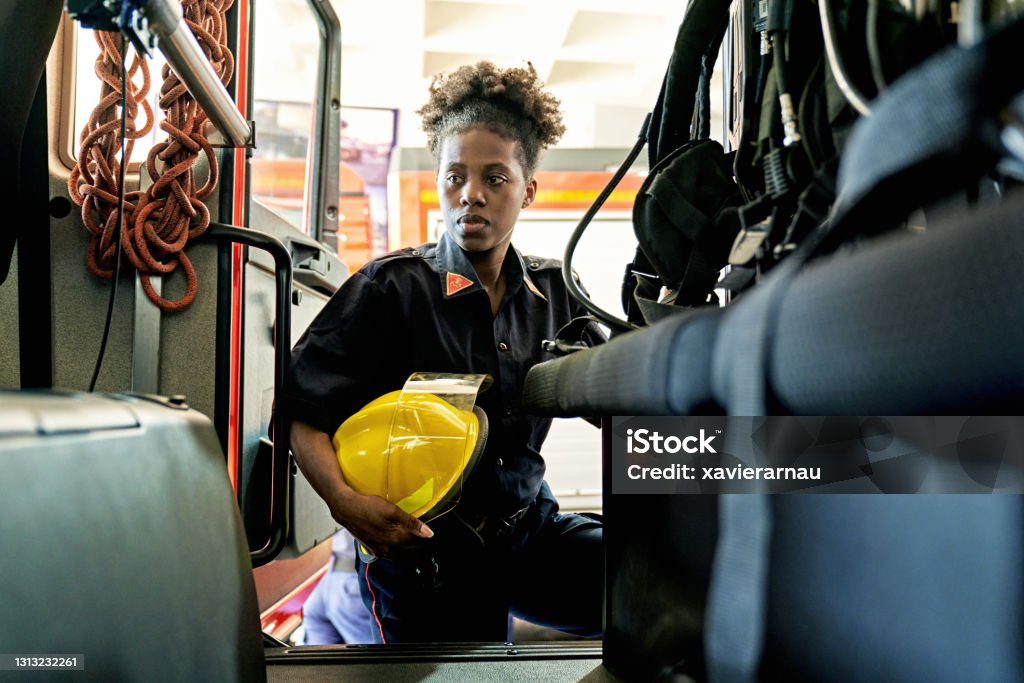 Black Female Firefighter with Helmet Boarding Fire Engine Early 30s woman in station wear with helmet stepping into vehicle and looking away from camera with serious expression. Firefighter Stock Photo