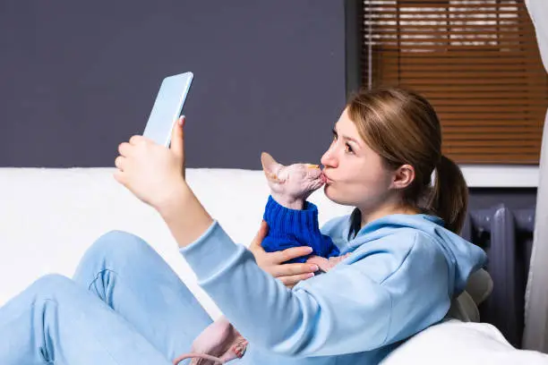 Photo of Loving girl taking selfie with a cat at home near the window. Cheerful girl kissing her pet in a domestic clothes and taking pictures with phone on a front camera. Technology lifestyle with pets